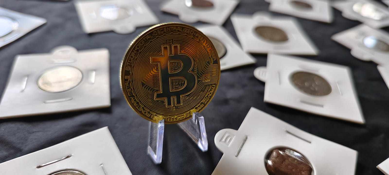 Close-up of a bitcoin coin standing among various currency coins in protective cases on a dark surface.
